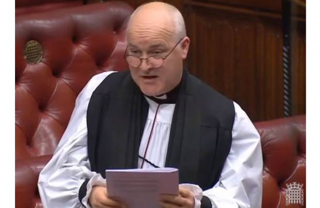 Head and shoulders of bishop in robes standing in front of red leather benches
